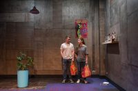 Two actors on stage holding shopping bags bottles on shelf