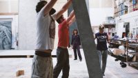 Scenic art students holding large backdrop, teacher in background