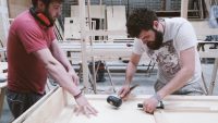 Two students constructing wooden pieces, one holding a hammer