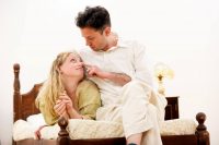 Female lying on bed looking up at male who is sat beside her with his finger on her nose