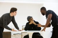 students in rehearsals in the Weston Studio - one male sits at a table with two males on either side talking to him