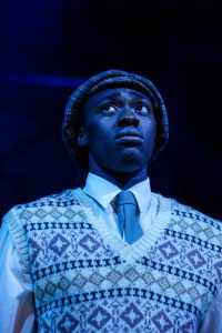 Male actor looking upwards wearing a patterned tanktop, tie and hat