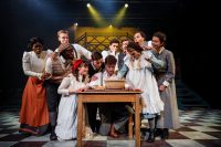 Group picture of eleven actors, all dressed in Edwardian costume, stood and knelt around a table which has a bowl and some bread on it