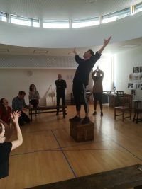 Student stood on box with arms outstretched being watched by a crowd in rehearsal