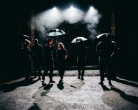 Figures holding umbrellas walking under bright lights and rain in The Laramie Project