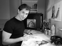 Black and white photo of design student drawing at a desk with a model set in the background