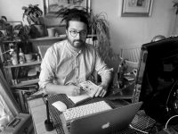 Black and whiite photo of student sat at desk holding a pen and drawing in a book, computers on desk