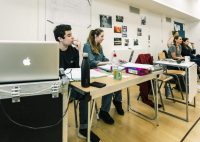 Two production arts students sat at table watching rehearsals. Teacher and other student sat at another table to their right watching rehearsals, and an acting student stood behind them