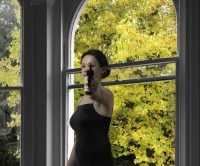 Young woman pointing gun at viewer, standing in front of an open window