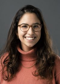 Headshot of female smiling at camera with dark brown hair, glasses and an orange roll neck jumper