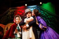 Male actor on stage, in Edwardian suit and hat, holding a baby, surrounded by two other actors in Edwardian dresses