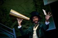 Actor on stage, in an Edwardian suit and hat, holding a megaphone and paper