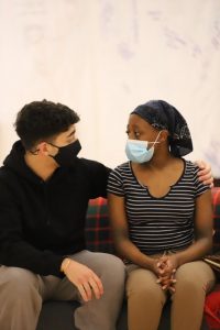 Two actors wearing masks, sat on sofa with arm aound the other