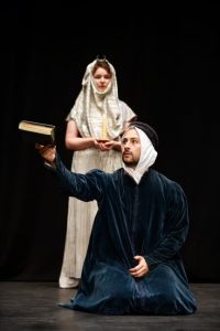 Actors dressed in robes, one knelt in foreground holding book, other stood behind