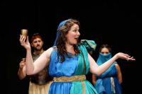 Female actor dressed in robes and headdress with arms outstretched, holding a cup