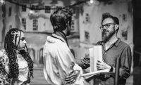 Black and white photo of a director speaking to two acting students in front of a theatre set