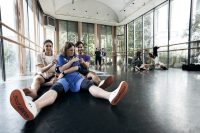 Three students are sat on the floor of a glass fronted studiowith different facial expressions